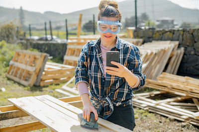 Woman in protective glasses text messaging on cellphone while sanding down wooden planks with orbit sander in countryside on summer day