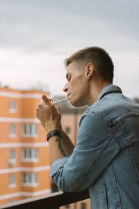 Side view of young man smoking cigarette while standing against sky in balcony