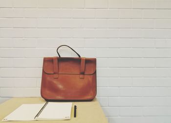 Bag and spiral notebook by pen on table against brick wall
