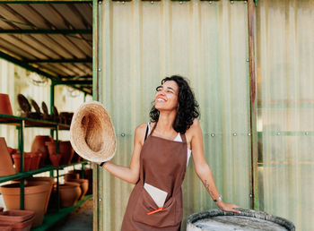 Smiling young woman holding hat while standing against wall