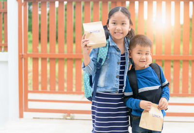 Girl with brother holding packed lunch against gate