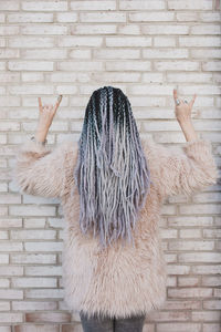 Rear view of woman wearing fur coat while gesturing horn sign against brick wall