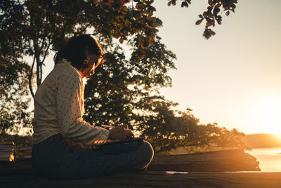 Man sitting by tree against sky during sunset