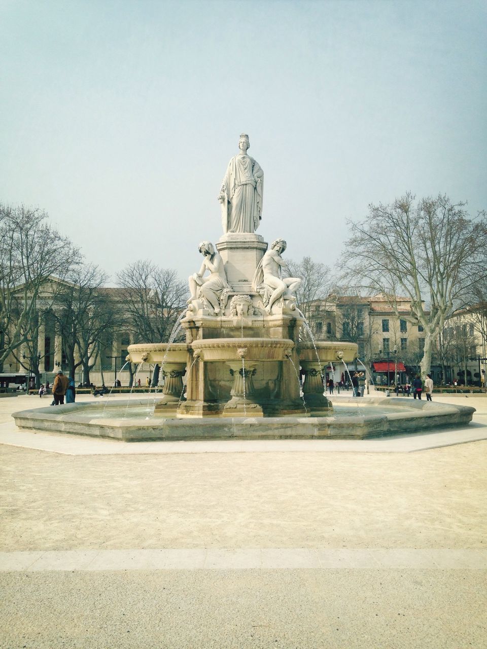 clear sky, tree, building exterior, architecture, built structure, incidental people, statue, sculpture, fountain, transportation, day, street, human representation, car, sky, travel, travel destinations, city, bare tree, outdoors