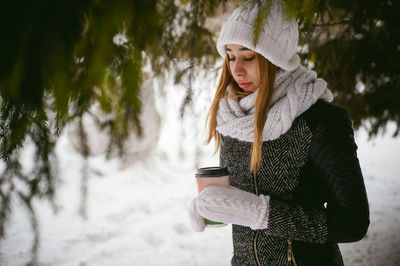 Young woman wearing warm clothing while holding disposable cup amidst trees during winter