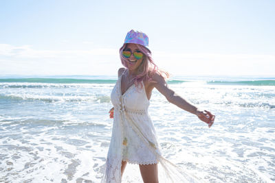 Content female with pink hair and in summer dress enjoying freedom with outstretched arms while standing on beach against sea and looking at camera