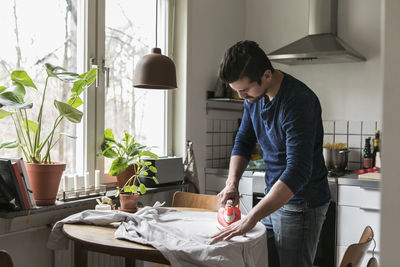 Young man ironing shirt on table by window while standing at kitchen