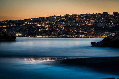 Illuminated city by sea against sky at sunset