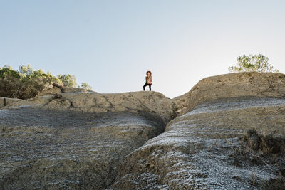 From below of female tourist in hat standing on rocky formation and looking away under cloudless sky in sunny day