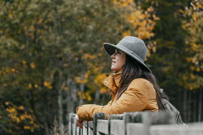 Side view portrait of young woman leaning on wooden fence by road in autumn woods