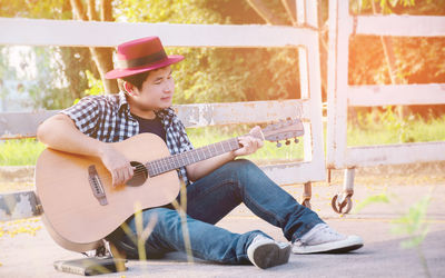 Full length of man playing guitar while sitting against fence