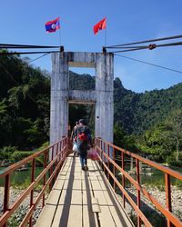 Rear view of person walking on footbridge over river against blue sky