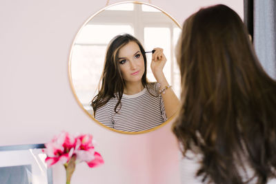 Girl with dark hair stands in front of a mirror and does makeup, holding a brush in her hand