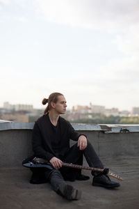 Full length of thoughtful young man with guitar sitting on building terrace