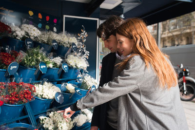Side view of happy young couple standing near stall with flowers and choosing bouquets while spending time together in city