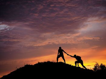 Silhouette man helping friend while climbing on mountain against cloudy sky during sunset