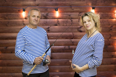 Portrait of man holding fishing rod by pregnant woman while standing against wall