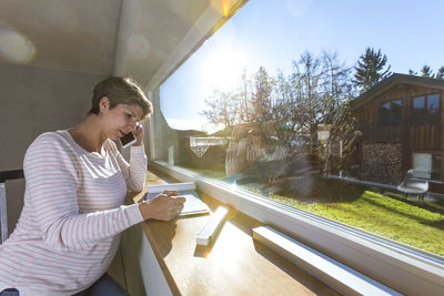 Pregnant woman talking on phone while reading document by window at home on sunny day