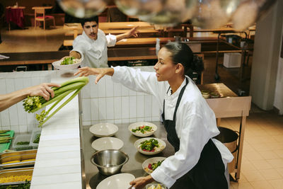 Female chef taking celery while male colleague passing salad bowl in commercial kitchen