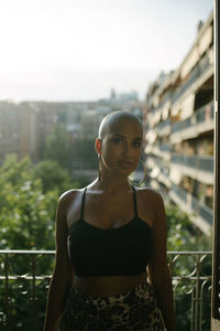 Black female with bald hairstyle and in trendy outfit standing on terrace of outdoors cafe and relaxing while looking at camera