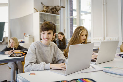 Happy boy looking away while sitting with laptop by female friend at desk in classroom