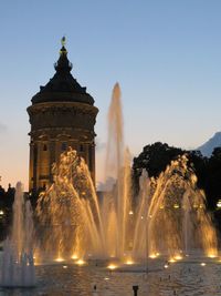 Water fountain in illuminated building against clear sky