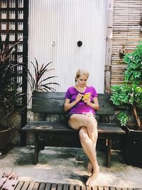 Woman using mobile phone while sitting on bench against wall