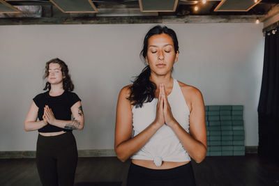 Calm slender ladies in activewear with namaste standing in mountain position on sports mats on wooden floor and meditating in spacious contemporary gym with closed eyes