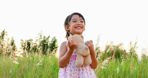 Happy girl holding teddy bear while standing on grassy field against clear sky