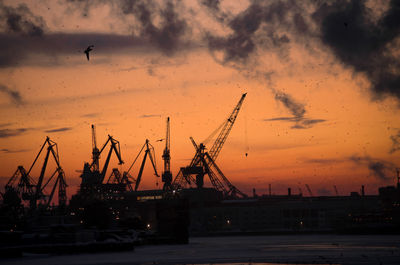 Silhouette cranes at construction site against sky during sunset