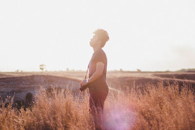 Side view of man with camera standing on field against clear sky