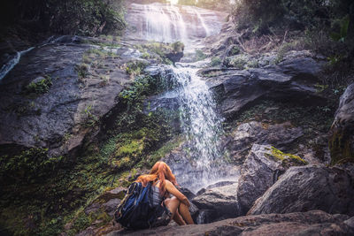 Rear view of woman with sitting on rock in forest against waterfall