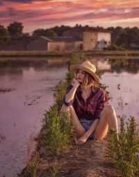 Side view of young woman sitting on field against sky during sunset