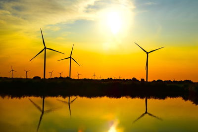 Silhouette wind turbines by lake against sky during sunset