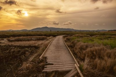 Dirt road leading towards field against sky during sunset