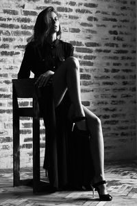 Full length of confident beautiful woman with hand on chin sitting on chair against brick wall
