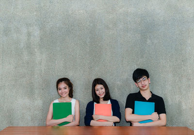 Portrait of friends holding book while sitting by table against wall in classroom