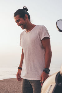 Happy adult ethnic male in casual t shirt standing next to motorbike at seaside at sunset time