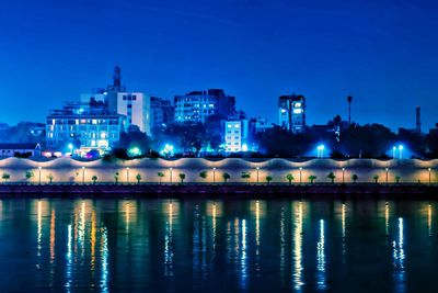 Illuminated buildings by lake against blue sky at night