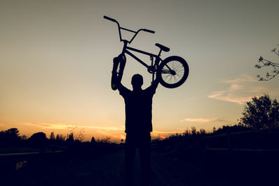 Rear view of man lifting bicycle against sky during sunset