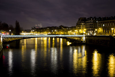 Illuminated bridge over river by buildings against sky at night