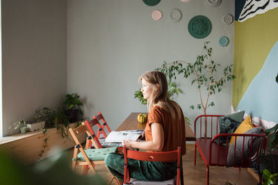 Woman sits at table with empty chairs, looks at window at home.