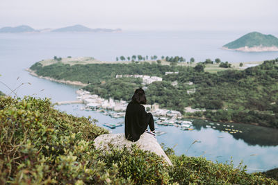 Rear view of woman sitting on rock by sea against sky