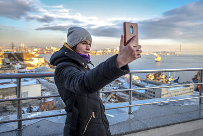 Man photographing while standing in city against sky during winter