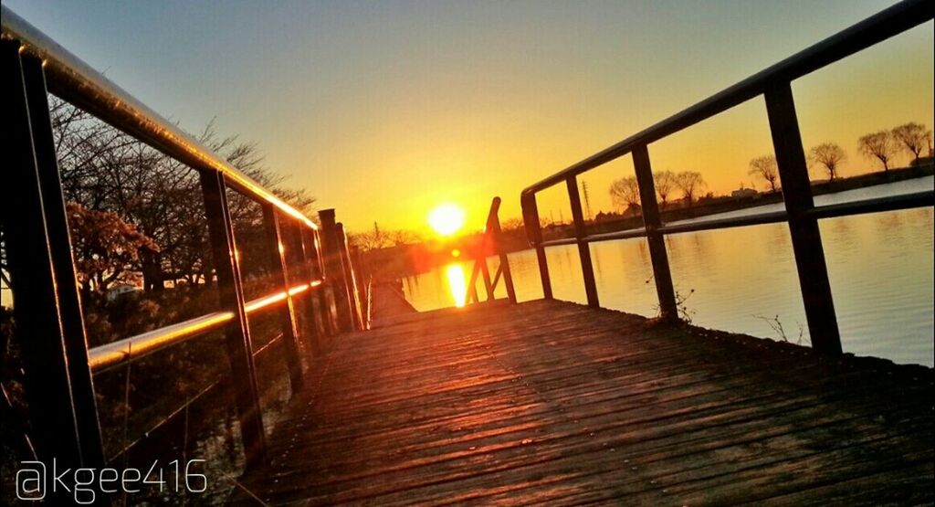 sunset, orange color, sun, railing, built structure, the way forward, sky, sunlight, transportation, water, architecture, diminishing perspective, nature, clear sky, lens flare, outdoors, tree, sunbeam, reflection, beauty in nature