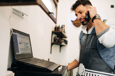 Crop bearded male barber speaking on wireless phone handset against netbook with open program on screen in beauty salon