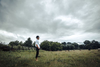 Side view of young man standing on grassy field against cloudy sky