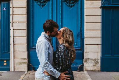 Side view of happy young couple in casual clothes hugging and kissing while standing against aged stone building with blue doors on city street