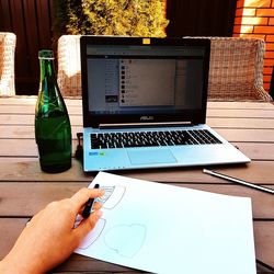 Cropped hand of person holding pen drawing on paper with laptop and beer on table