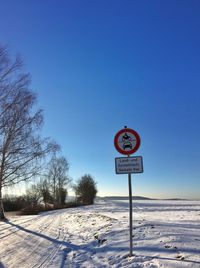 Metallic sign by snow covered road against clear blue sky